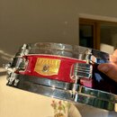 Červený snare drum s chromovanými obručmi a mechanismy. Na korpusu je zlatá štítková nálepka s nápisy TAMA, SNARE DRUM a SERIAL No. 210024, pod ní je…