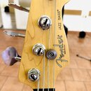 Detail hlavy basové kytary z přírodního dřeva. Na dřevě je černý nápis „Fender“, pod ním text „MADE IN USA“ a vedle něj svisle nápis „JAZZ BASS®“. Ná…