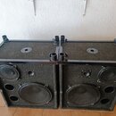 Dvě černé reproduktorové bedny s povrchem z tmavého kobercového materiálu a kovovými hranami. Každá bedna obsahuje jeden velký a jeden menší reproduk…