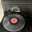 Černý gramofon s umístěnou vinylovou deskou. Na středu desky je červenobílé kulaté logo s motivem stylizovaného symbolu. V pravé části je vidět ramen…