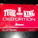 Červená kartonová krabice s nápisy. Na horní straně je bílý text TUBE KING DISTORTION, logo Ibanez a grafika znázornující postavu s korunou a nápisem…