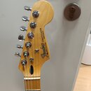 Fotografie zachycuje hlavu elektrické kytary z přírodního světlého dřeva s chromovanými ladičkami. Na hlavě je černý nápis Fender® STRATOCASTER MADE…