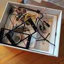 Bílá krabička s vnitřním lůžkem obsahuje rozložené elektronické součástky, potenciometry a barevné vodiče. Jsou zde dvě černé kovové rámy s otvory a…