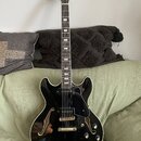 Černá polodublová elektrická kytara s lesklým povrchem a f-otvory. Na hlavě hmatníku je zlatý nápis "Joe Pass". Nástroj má zlaté ladičky, černý pickg…