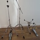 Sada chromovaných kovových stojanů a pedálů. V popředí jsou dva stojany s trojnožkou a jeden hi-hat stojan, vpravo je umístěn basový pedál. Pedály ma…