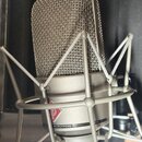 Kondenzátorový mikrofon v šedé barvě s kovovou síťkovou hlavicí, umístěný v odpružíného držáku (shock mount). Na těle mikrofonu je viditelné červené…