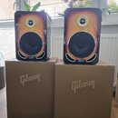Dva studiové monitory s lesklým povrchem v přechodové barvě od žluté po černou, umístěné na dvou hnědých kartonových krabicích. Na přední straně každ…
