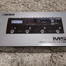 Krabice s efektem Boss MS-3 Multi Effects Switcher v šedé barvě. Na horní části je vidět ovládací panel s displejem, na kterém je nápis BOSS MS-3, a…
