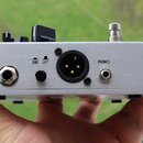 Stříbrné kovové zařízení s broušeným povrchem. Na zadní straně jsou nápisy GND a LIFT u přepínače, konektor XLR a výstup s nápisem PHONES. V levém do…