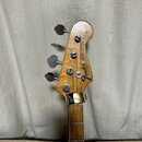 Fotografie zachycuje hlavu basové kytary z přírodního dřeva se stříbrnými ladičkami. Na hlavě je černý nápis Fender PRECISION BASS a pod ním menší te…