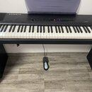 Černý digitální piano s bílými a černými klávesami, umístěné na černém stojanu. Na levé straně horního panelu je bílý nápis KURZWEIL a vpravo nápis M…