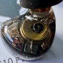 Detailní pohled na sluchátko v průhledném plastovém krytu. Uvnitř jsou vidět technické komponenty, včetně zlatavého kruhového elementu. Na spodní čás…