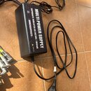 Černý obdélníkový zdroj napájení s několika výstupními kabely. Na horní straně je bílý nápis MULTI POWER SUPPLY, SEPARATE ISOLATED 8X DC10V / 100mA a…
