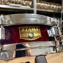 Detailní pohled na červený snare drum s chromovanými obručemi. Na korpusu je zlatý štítek s nápisy TAMA, SNARE DRUM a MADE IN JAPAN. Na štítku je dál…