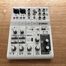 Bílá mixovací konzole značky YAMAHA, model AG06 MIXING CONSOLE. Zařízení obsahuje různé vstupy a výstupy, včetně konektorů XLR, jack 6,35 mm a USB. N…