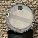 Fotografie zobrazuje snare drum (malý buben) s bílou blánou. Na bláně je černé logo MAPEX v podobě štítu. V dolní části blány je nápis UT REMO COATED…