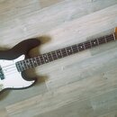 Černá elektrická basová kytara s bílým pickguardem a světle hnědým hmatníkem. Na hlavě nástroje je viditelné logo Squier a nápis by Fender. Instrumen…