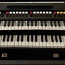 Černý dvoumanuálový synthesizér s dřevěnými bočními panely. Horní panel obsahuje řadu barevných tlačítek a posuvníků, vpravo na spodním panelu je náp…