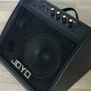 Černý reproduktor s kovovou mřížkou, na které je umístěno stříbrné logo JOYO. Horní ovládací panel obsahuje červený vypínač POWER, čtyři otočné knofl…