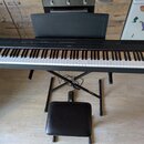 Černý digitální piano s klávesnicí, umístěné na černém X-stojanu s přiloženou černé polstrovanou židličkou. Na horní liště nástroje je uprostřed bílý…