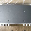 Černý obdélníkový elektronický přístroj v polystyrenovém balení. Horní strana je hladká s osmi kulatými prohlubněmi v rozích a po stranách. V dolní č…