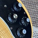 Detailní záběr na ovládací prvky kytary s černým pickguardem a dřevěným tělem. Jsou zde tři černé oválné knoflíky s kovovými zrcadlivými vršky, dva z…