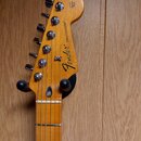 Fotografie zachycuje hlavu a část hmatníku elektrické kytary z dřeva s žlutohnědou povrchovou úpravou. Na hlavě je černý nápis Fender® STRATOCASTER®.…