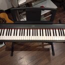 Černý digitální klavír značky YAMAHA umístěný na černém stojanu. Nástroj má bílé a černé klávesy s červeným pruhem nad nimi. V horní části je umístěn…