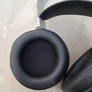 Černé sluchátka s šedým kovovým ná headbandem s perforací. Jsou vidět dva ušní polštáře z černé syntetické kůže s látkovým středem. Na fotografii nej…