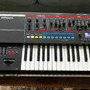 Černý syntetizátor s bílými a černými klávesami. Na levé straně je bílé logo Roland, v horní části jsou ovládací prvky s nápisy I-ARPEGGIO, LFO, OSC,…