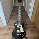 Černá elektrická kytara s tvarem singlecut a krémovým pickguardem umístěná na černém stojanu. Na hlavě hmatníku je bílé logo „Epiphone“. Nástroj má d…