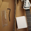 V kartonové krabici s polystyrenovými výplněmi je vidět hlava a hриф elektrické kytary bílé barvy. Na hlavě je černý nápis Schecter a pod ním menší t…