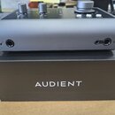 Šedý audio rozhraní s matným povrchem umístěný na černé krabici. Na přední straně přístroje jsou dva menší a jeden velký ovládací knoflík, vstupní ja…