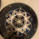 Kulatý kovový nástroj typu handpan v tmavě modré barvě. Povrch je zdoben bílo-stříbrným geometrickým vzorem připomínajícím květ života. Na horní část…
