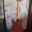 Zadní pohled na elektrickou kytaru v červenobronzové barvě, umístěnou na černém stojanu. Na zadní straně těla je vidět černé logo s nápisem "Squier".…