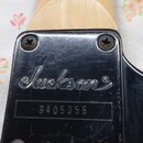 Detail zadní strany hlavy kytary s kovovým černým krytem. Na krytu je vyryto logo Jackson a sériové číslo 9405355. Povrch krytu vykazuje drobné škráb…