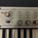 Detailní pohled na ovládací panel stříbrného klávesového nástroje. Panel obsahuje nápisy ORIGINAL VALUE, TIMBRE SELECT a tabulku s funkcemi ASSIGN, A…