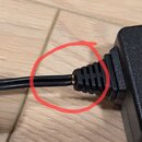 Detailní záběr na připojení černého kabelu k černé plastové krabičce napájecího adaptéru. V místě vstupu kabelu do odlehčovacího prvku je viditelné p…