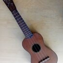 Hnědý dřevěný ukulele s tmavým hmatníkem. Nástroj vykazuje známky opotřebení, jsou vidět škrábance a nečistoty na povrchu těla. Na hlavě jsou struny…