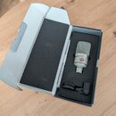 V otevřené krabici s černou pěnovou vložkou je umístěn stříbrný kondenzátorový mikrofon a černý držák. Na těle mikrofonu je červené logo v kosočtverc…