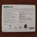 Fotografie znázorňuje zadní stranu bílého obalu od mikrofonu s nápisem RØDELink™ LAV. Text v několika jazycích popisuje vlastnosti produktu a sekce B…