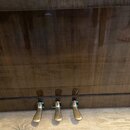 Detail spodní části pianina s tmavým, vysoce lesklým dřevěným povrchem. V dolní části jsou vidět tři mosazné pedály. Povrch nástroje vykazuje odrazy…
