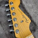 Fotografie zachycuje hlavu elektrické kytary z jasného dřeva s šestnácti chromovanými ladičkami. Na povrchu je černý nápis "Fender STRATOCASTER". Pov…