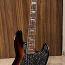 Elektrická basová kytara s trupem v barevnosti sunburst a černým pickguardem s texturou. Na hlavě hmatníku je nápis Fender Jazz Bass. Nástroj vykazuj…