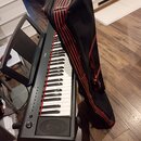 Černý digitální klavír na černém stojanu s nápisem YAMAHA na ovládacím panelu. Vedle nástroje je položen černý transportní vak s červenými svislými p…
