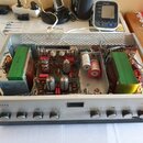 Stříbrný elektronický přístroj s otevřeným šasi, ve kterém jsou vidět dva velké zelené transformátory, kondenzátory a elektronky. Na předním panelu j…
