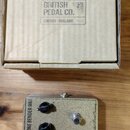 Na fotografii je hnědý kartonový krabic s nápisem BRITISH PEDAL CO. LONDON - ENGLAND. Před krabicí leží efekt pedál v béžovém kovovém šasi s textem T…