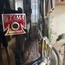 Detail černé lesklé bubnové skořápky s kovovými lugy. Na korpusu je nalepený červeno-stříbrný štítek s nápisy TAMA SWINGSTAR, No. 84-14027 a MADE IN…