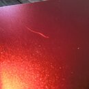 Detailní záběr na červený lakovaný povrch hudebního nástroje s metalickým efektem. V horní části je vidět podélný škrábanec v laku. V pravém okraji j…