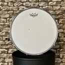 Bílá bubnová blana s kovovým obručem. V horní části je černý nápis REMO, pod kterým jsou menšími písmeny uvedeny označení AMBASSADOR, CLEAR a MADE IN…